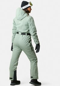 Kobiecy kombinezon narciarski w jasnym zielonym kolorze, z kapturem, czarnym paskiem, zapinanymi kieszeniami oraz czarnymi akcentami przy mankietach i nogawkach. Miła w dotyku tkanina.