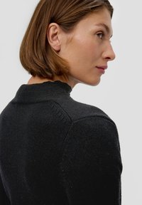 Zwarte gebreide trui met een hoge halslijn, voorzien van een gestructureerde afwerking en subtiele stikdetails op de schouders, zijaanzicht weergegeven.