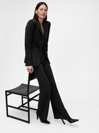 Costume noir sur mesure avec un blazer ajusté et un pantalon évasé. Porté avec des talons pointus noirs, assis sur une chaise noire simple.
