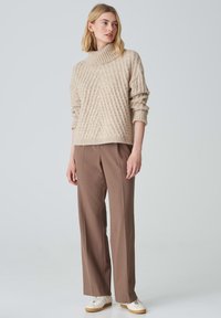 Beige gestrickter Pullover mit hohem Kragen und gerippter Textur, kombiniert mit braunen maßgeschneiderten Hosen und weißen Sneakers. Einfacher, lässiger Schnitt.