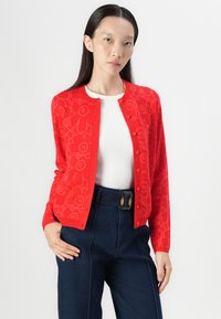 Red cardigan with floral pattern, round neckline, and button closure, worn over a white top, paired with dark blue denim trousers.