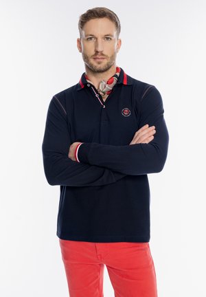 Mężczyzna z założonymi skrzyżowanymi ramionami, ubrany w granatową koszulkę polo z długim rękawem, wzorzysty szalik na szyi i czerwone spodnie, stojący na jednolitym białym tle.