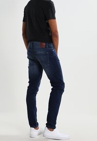 Mörkblå denimjeans med slim fit, med en brun läderlapp på bakre midjebandet och två bakfickor. Bärs med en svart skjorta och vita sneakers.