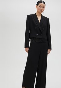 Svart dubbelknäppt kavaj med fransdetaljer vid fållen, tillsammans med vida svarta byxor. Slät tyg, strukturerad design, modern silhuett.