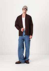 Brun, lett jakke med en avslappet passform, kombinert med løstsittende blå cargo-jeans. Brune sko og en beige caps fullfører stilen.