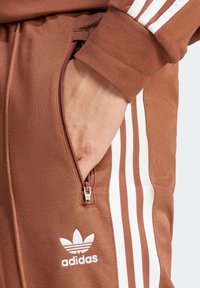 Bruna atletiska joggers med vita sidosträck, blixtlåsficka och broderad Adidas-logga. Tillverkade av texturerat tyg.