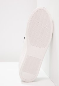 Semelle de chaussure blanche avec motif de grip texturé et détails de la marque en relief. Matériau lisse et caoutchouteux avec des bords arrondis et un design minimaliste.