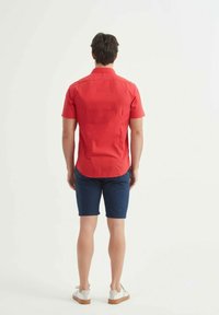 Chemise rouge à manches courtes, shorts bleus et chaussures blanches. La chemise a une coupe droite et une texture uniforme, portée de dos.
