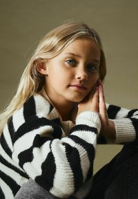 Sweter dziany w czarno-białe paski z teksturowanej tkaniny; luźny krój, długie rękawy oraz szeroki kołnierz. Modelka ma jasnoblond włosy.