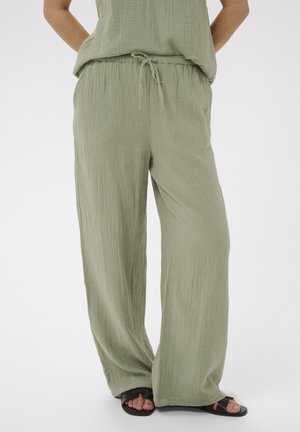 Luźne, jasnozielone spodnie o fakturowanej powierzchni ze sznurkiem w talii, noszone z czarnymi, odkrytymi sandałami, ukazujące część dopasowanego topu i skrzyżowane nogi.