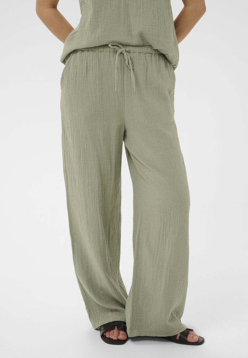 Pantalon ample vert clair texturé avec taille à cordon porté avec des sandales noires à bout ouvert, montrant une partie d'un haut assorti et des jambes croisées.