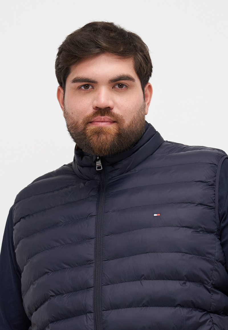 Gilet matelassé bleu marine avec motif de couture horizontal, col montant et fermeture éclair à l'avant. Petit logo sur la poitrine gauche. Tissu doux et isolant.