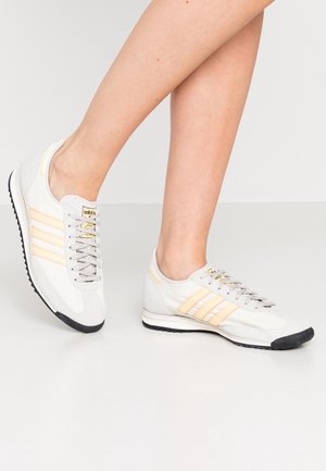 Personne portant des baskets Adidas blanches et beiges avec des semelles noires, debout sur un sol blanc devant un fond uni.