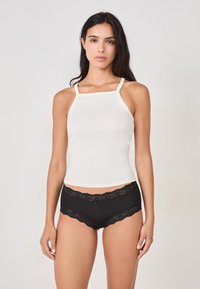 Femme aux longs cheveux noirs portant un haut sans manches blanc à côtes et une culotte en coton noire bordée de dentelle, se tenant devant un fond uni.