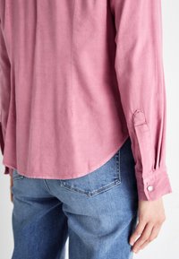 Chemise rose pâle à manches longues avec un ourlet incurvé, des poignets à boutons et une texture douce, associée à un jean bleu avec des poches arrière.
