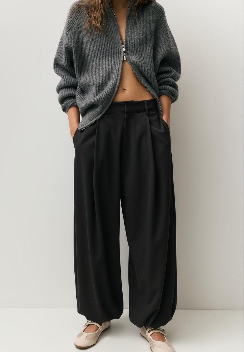 Személy szürke cipzáras pulóverben, bő, fekete, széles szárú nadrágban és bézs balerinacipőben, kezét a zsebében tartva, egyszerű világos háttér előtt áll.