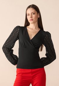 Haut noir avec un décolleté en V profond et un corsage ajusté, présentant des manches bouffantes transparentes et texturées avec un motif géométrique. Assorti à un pantalon rouge.