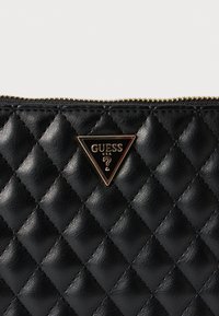 Czarna pikowana skórzana kosmetyczka z zamkiem błyskawicznym w złotym odcieniu. Z przodu znajduje się trójkątny detal z logo oraz wytłoczonym tekstem. Powierzchnia o fakturze.