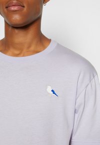 Light purple cotton t-shirt featuring a white and blue embroidered bird design on the left chest. Crew neck and short sleeves.