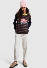 Duga majica s prugama u smeđoj i crnoj boji s grafikonom Hogwartsa, uz krem pantalone i smeđe planinarske cipele, a na glavi ružičasta kapa.