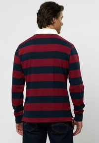 Férfi háttal, hosszú ujjú rögbi pólót viselve, amely széles, vízszintes bordó és tengerészkék csíkokkal, valamint fehér gallérral van díszítve.