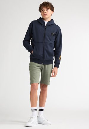 Donkerblauwe hoodie met ritssluiting en een logopatch, kangoeroezakken, gecombineerd met olijfgroene shorts. Model draagt witte sneakers en gestreepte sokken.
