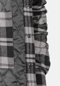 Steppjacke mit einem schwarz-grauen Karomuster, einem zusätzlichen grauen Stoffabschnitt und metallenen Druckknöpfen auf der Vorderseite.