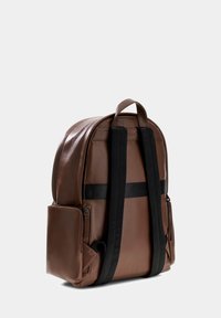 Sac à dos en cuir marron avec une texture lisse, des bretelles noires réglables et une poche zippée sur le côté. Présente une forme arrondie et un design minimaliste.