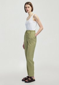 Lehký bílý top bez rukávů v kombinaci s kalhotami s vysokým pasem v olivově zelené barvě, které mají zavazovací pas, boční kapsy a tmavě hnědé sandály.