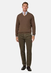 Rudas v-kaklo megztinis ant šviesiai mėlynos marškinės, derinamas su alyvuogių žalumo štramo džinsų kelnėmis ir juodais elegantiškais batais. Neutrali aplinka.