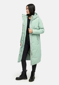 Cappotto lungo imbottito verde menta con cappuccio, chiusura a bottoni a pressione e tasche laterali, abbinato a jeans neri e stivali militari.