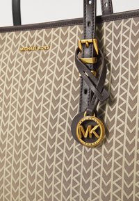 Brun tote bag med geometrisk tan-mønster, guld logo-tag og mørkebrune remme. Har en subtil tekstur og metalliske accenter.