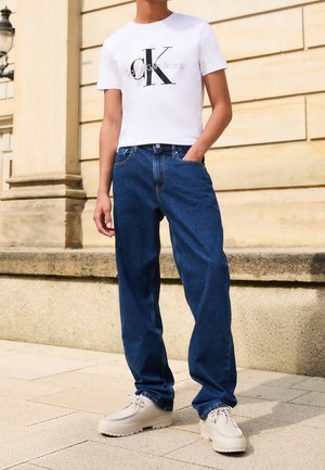 Person iført hvid Calvin Klein logo t-shirt, blå jeans og beige snørestøvler stående udendørs ved en beige stenmur.