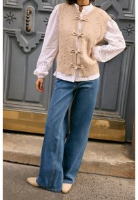 Beige strikket vest med knapdetaljer over en hvid skjorte, kombineret med flagrende blå jeans og beige sko, sat op mod en dekorativ dør.