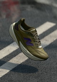 Oliivroheline jooksujalats, millel on lilla logo, koos võrgust pealmise osa, lamedate paelte ja paksu valge tallaga. Kerge disain jooksmiseks.