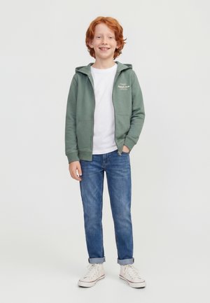 Garçon roux souriant portant un sweat à capuche vert zippé, un t-shirt blanc, un jean bleu retroussé et des baskets blanches, debout sur un fond blanc.