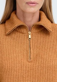 Rostfärgad tröja med ribbat mönster, som har en dragkedja vid halsen och ett mjukt, stickat tyg. Gula dragkedjespännen är synliga.