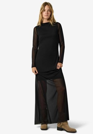 Une femme porte une robe longue noire à manches transparentes et à pans de jupe, avec des bottines beige clair, sur fond blanc.