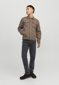 Giacca di jeans marrone con tasche frontali, bottoni argentati e colletto, abbinata a jeans grigio scuro e calzature nere. Sfondo uniforme.