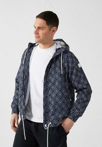 Uomo che indossa una giacca con cappuccio blu navy a motivo sopra una t-shirt bianca e pantaloni neri, guarda di lato con le mani nelle tasche.