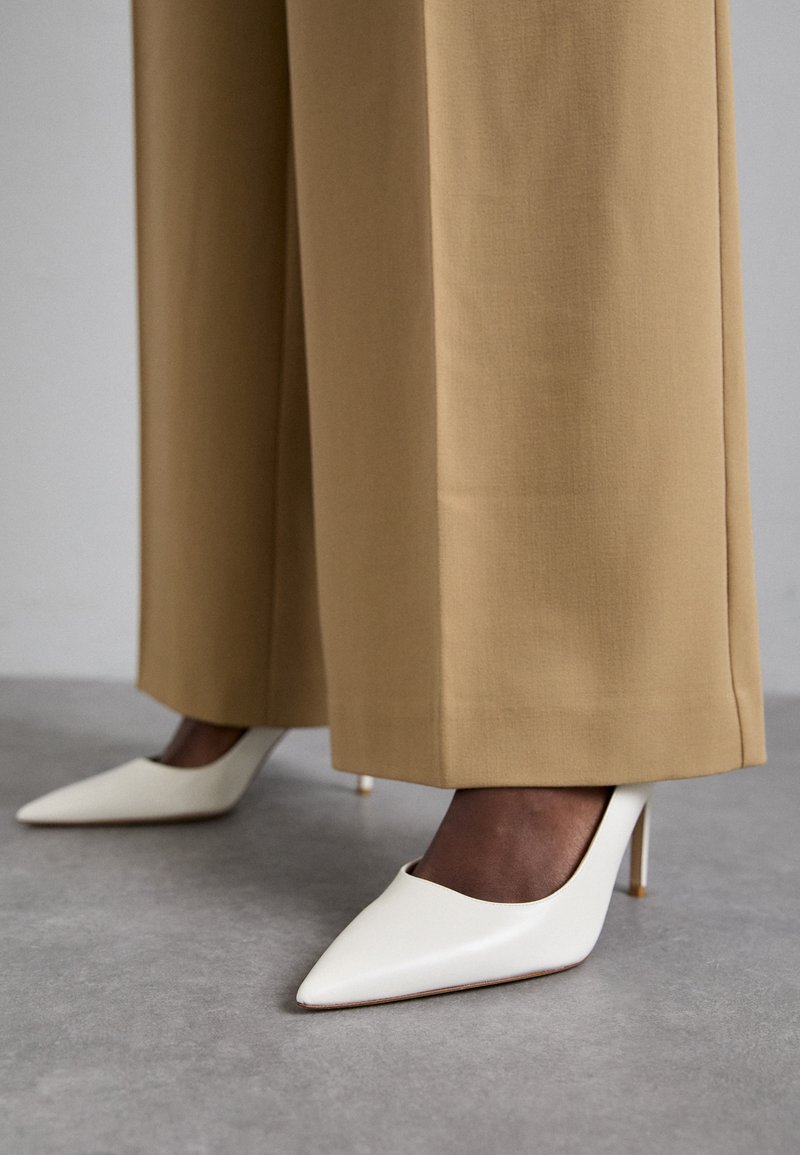 White pointed-toe heels with a glossy finish, paired with wide, beige trousers. The heels have a slim, golden stiletto.