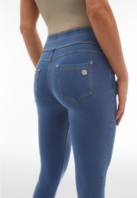 Primer plano de una persona que lleva jeans de mezclilla azul elástica de cintura alta con bolsillos traseros y una camiseta beige, de pie con una mano relajada.