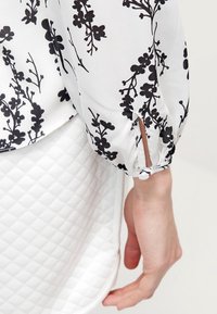 Vit blus med svart blommönster, med puffärmar och en knäppt slitsdetalj. Slät tyg mot en texturerad vit kjol.