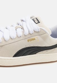 Baskets Puma Suede XL beige avec des lacets blancs épais, bande noire Puma, logo doré et semelle texturée couleur blanc cassé.