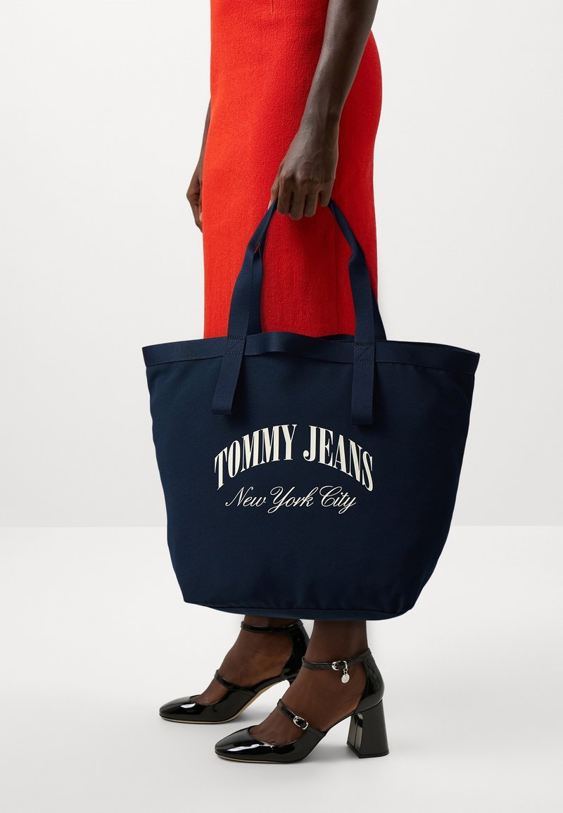 Personne en robe rouge tenant un grand sac cabas bleu marine avec le texte "Tommy Jeans New York City", portant des chaussures noires à talons blocs vernies.