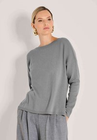 Frau in grauem Strickpullover und grauen plissierten Hosen, accessoriert mit großen goldenen Hoops, steht vor einem beigen Hintergrund.