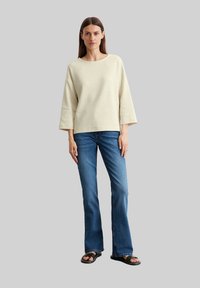 Haut à rayures beige à manches longues, coupe décontractée, avec des manches larges, associé à un jean bleu évasé et des sandales noires, se tenant devant un fond gris.