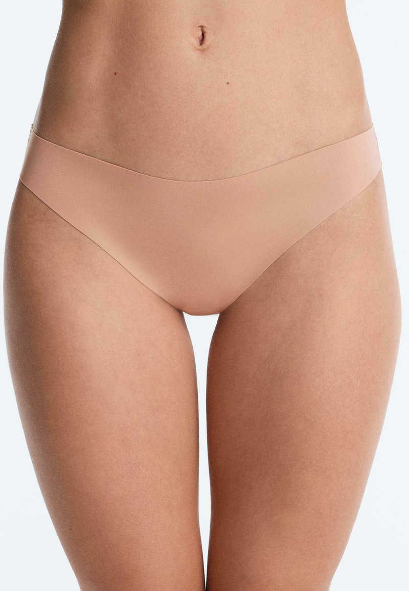 Femme portant des sous-vêtements style bikini couleur nude sans coutures, montrant le bas de l'abdomen et le haut des cuisses sur un fond blanc uni.