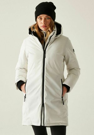 Donna che indossa un cappotto invernale bianco con cappuccio e dettagli con cerniere nere, un berretto nero, mani nelle tasche, in piedi di fronte.