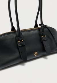 Μαύρη δερμάτινη τσάντα χειρός με δύο μακριές λαβές, κλείσιμο με χρυσό φερμουάρ και ανάγλυφο λογότυπο. Διαθέτει δομημένο σχήμα και ελάχιστες ραφές.